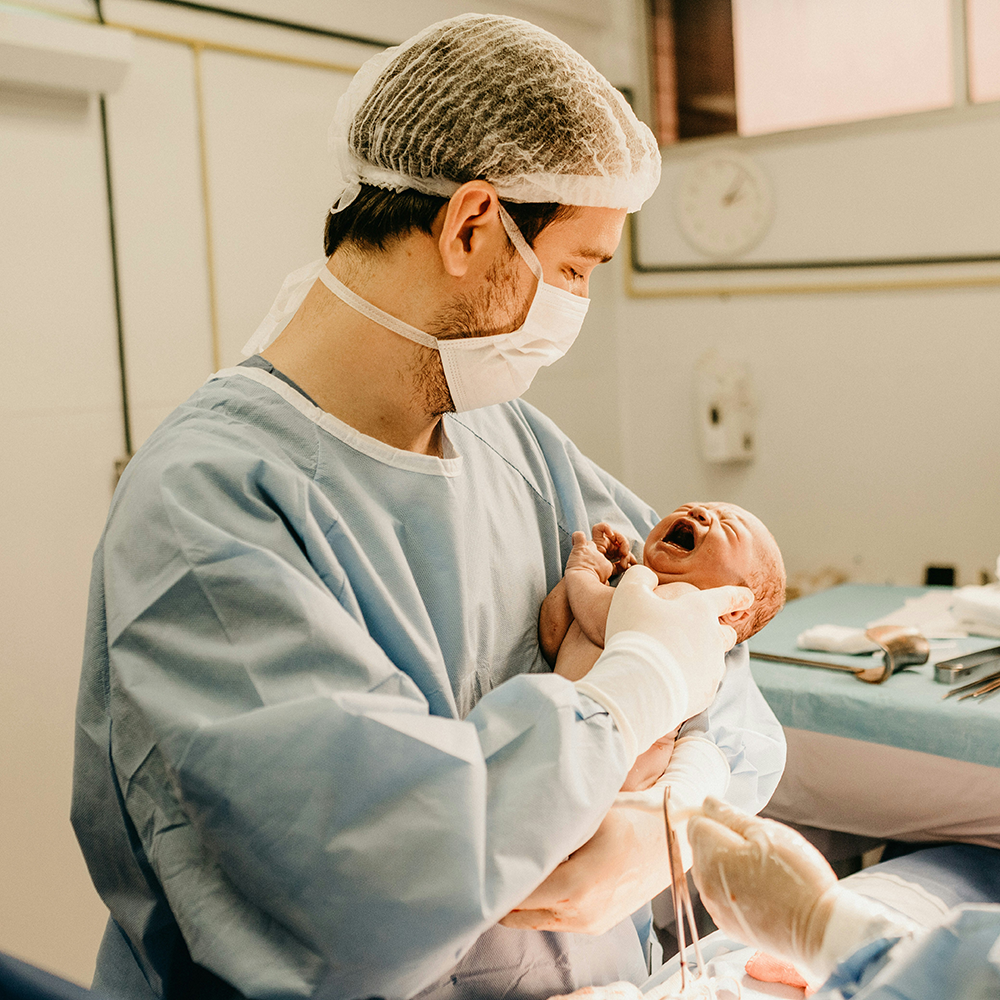 A healthcare worker in surgical attire holds a crying newborn in a hospital setting.